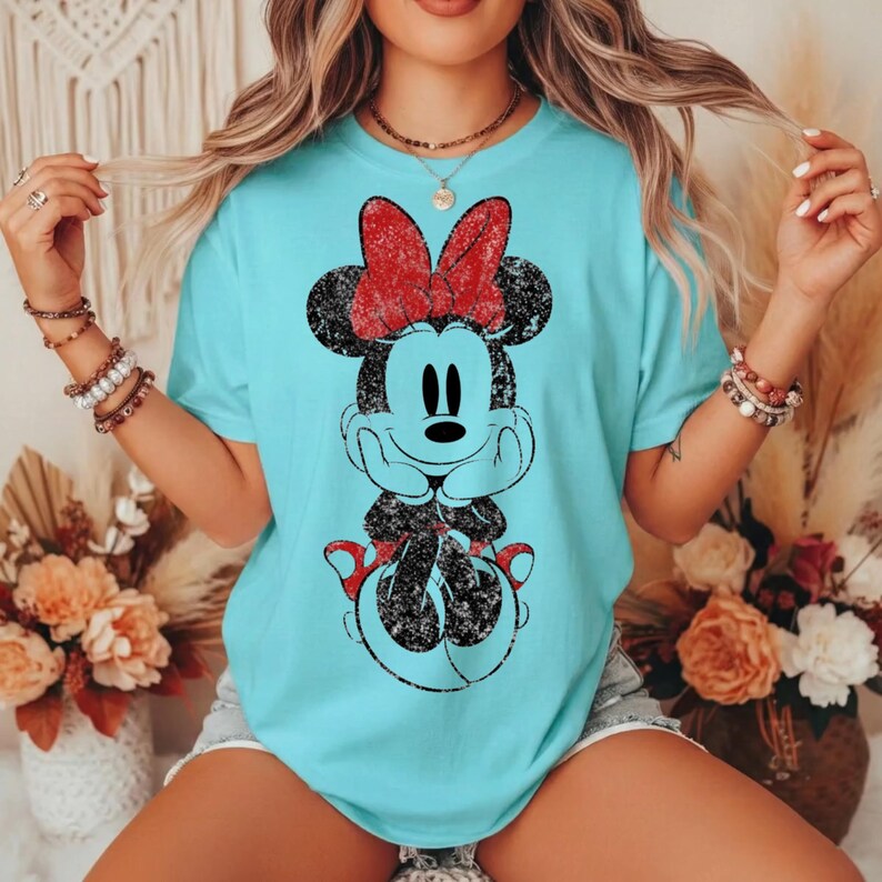 May include: A light blue t-shirt featuring a distressed graphic of Minnie Mouse with a red bow. The cartoon character is in a seated pose with hands under her chin. The shirt is a crew neck and short-sleeved.