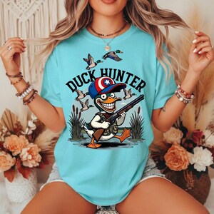 Op de afbeelding: Lichtblauw T-shirt met een cartoon eendenjager-ontwerp. De afbeelding toont een eend met een camouflagevest en een hoed met een ster, die een geweer vasthoudt. De woorden "DUCK HUNTER" staan boven de eend, met vliegende eenden op de achtergrond.
