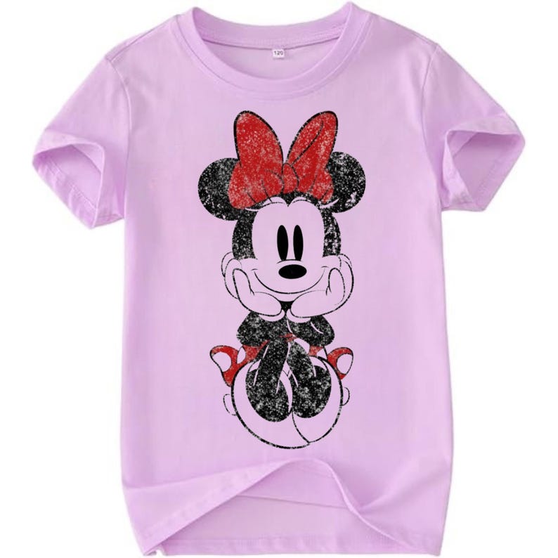 May include: A light purple t-shirt featuring a distressed graphic of Minnie Mouse. The cartoon character has a red bow, black ears, and a black and white outfit. The shirt has short sleeves and a crew neck.