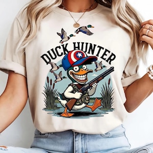 Op de afbeelding: Beige T-shirt met een cartoon eendenjager ontwerp. De afbeelding toont een eend met een geweer, een hoed met een ster embleem en de tekst "DUCK HUNTER". Vliegende eenden en riet zijn op de achtergrond.