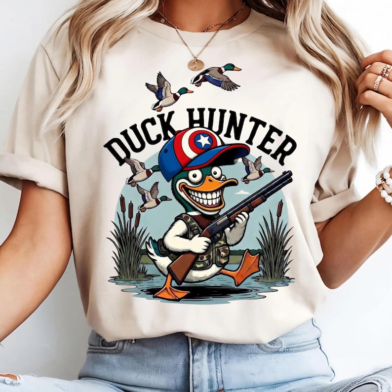 Op de afbeelding: Beige T-shirt met een cartoon eendenjager ontwerp. De afbeelding toont een eend met een geweer, een hoed met een ster embleem en de tekst "DUCK HUNTER". Vliegende eenden en riet zijn op de achtergrond.