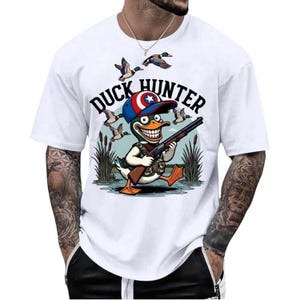 Op de afbeelding: Wit T-shirt met een cartoon eendenjager. De eend draagt een hoed en houdt een geweer vast, met de tekst "DUCK HUNTER" erboven. Eenden vliegen op de achtergrond.
