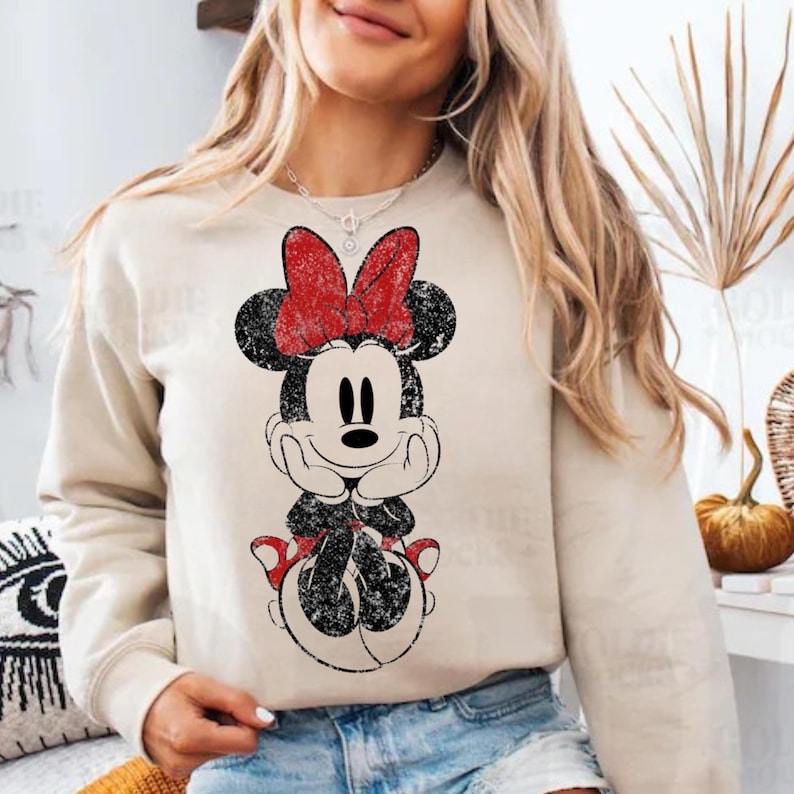 May include: Beige sweatshirt featuring a distressed graphic of Minnie Mouse with a red bow. The cartoon character is in black and white with red accents. The sweatshirt has long sleeves and a crew neck.