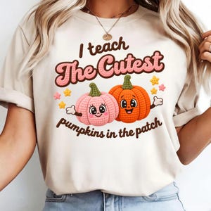 Peut inclure: Un t-shirt couleur crème avec un motif de deux citrouilles de dessin animé avec des visages, une rose et une orange, entourées de petites étoiles jaunes et roses. Le texte indique "I teach The Cutest pumpkins in the patch" en lettres marron et rose.