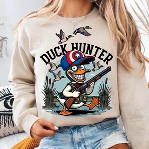 Op de afbeelding: Beige sweatshirt met een cartoon eendenjager-afbeelding. De afbeelding toont een eend met een hoed en een geweer, met de woorden "Duck Hunter" erboven. Eenden vliegen op de achtergrond.