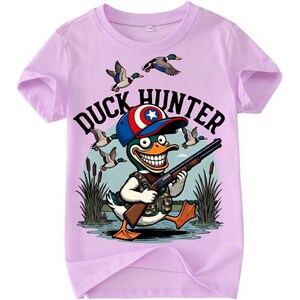 Op de afbeelding: Lichtpaarse t-shirt met een cartoon eendenjager met een geweer. De eend draagt een camouflagevest en een blauwe, rode en witte hoed. De tekst "DUCK HUNTER" staat boven de eend, met vliegende eenden op de achtergrond.