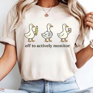 Peut inclure: T-shirt beige avec un motif de trois canards de dessin animé. Les canards tiennent un crayon, portent un t-shirt avec l'inscription "You Got This" et tiennent une tasse de café. Le texte "off to actively monitor" est en dessous.