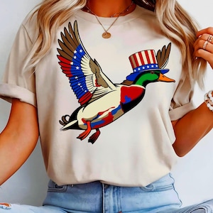 Peut inclure: T-shirt beige avec un dessin de canard de dessin animé. Le canard a des ailes rouges, blanches et bleues, et porte un chapeau haut de forme avec des étoiles et des rayures. Le canard est représenté en plein vol, ailes déployées. Design patriotique et fantaisiste.