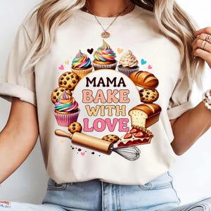 Peut inclure: T-shirt beige avec un motif coloré de pâtisseries, notamment des cupcakes, des biscuits et du pain. Le texte "MAMA BAKE WITH LOVE" est présenté dans un cercle. Le motif comprend également un rouleau à pâtisserie, des cœurs et une tarte aux fraises.