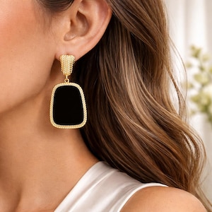 May include: Gold-toned rectangular drop earrings with a black center and a textured border. The earrings have a rectangular shape and a post-back closure. The earrings are shown on a person's ear.