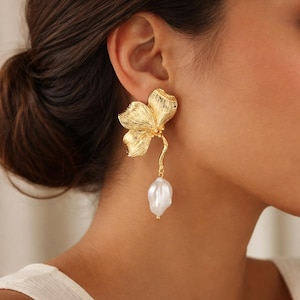 May include: A gold-colored floral earring with a dangling white pearl. The earring features a textured flower design with a curved stem leading to the pearl. The jewelry is worn on the ear.