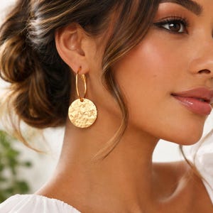 May include: Gold-coloured hoop earrings with a textured, hammered-metal disc pendant. The earrings are round and hang from the earlobe. The disc pendant has a hammered texture, catching the light. The earrings are a warm gold colour.