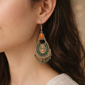 Könnte beinhalten: Vergoldete, verzierte Ohrringe mit orangefarbenen und grünen Akzenten. Die Ohrringe haben oben einen tropfenförmigen orangefarbenen Stein, in der Mitte ein dekoratives Design und kleine grüne und orangefarbene Perlen, die von unten hängen. Die Ohrringe sind zum Hängen gedacht.