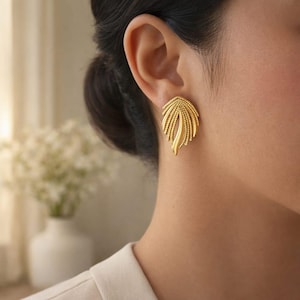 Puede incluir: Primer plano de un pendiente dorado en forma de hoja. El pendiente tiene una superficie texturizada con un diseño detallado. El pendiente se lleva en la oreja de una persona.