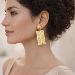 May include: Gold-toned earrings featuring a textured rectangular drop and a round stud. The earrings have a detailed, embossed pattern and a polished finish. These statement earrings are a bold accessory for any outfit.