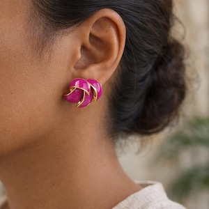 Può includere: Primo piano di un paio di orecchini color magenta con dettagli dorati. Gli orecchini hanno un design curvo unico. Gli orecchini sono sull'orecchio di una persona.