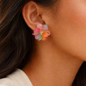 Peut inclure: Gros plan sur une boucle d'oreille à clou florale colorée. La boucle d'oreille présente un groupe de petits pétales translucides dans les tons de rose, d'orange, de vert et de bleu. La boucle d'oreille est portée sur l'oreille d'une personne aux cheveux foncés.