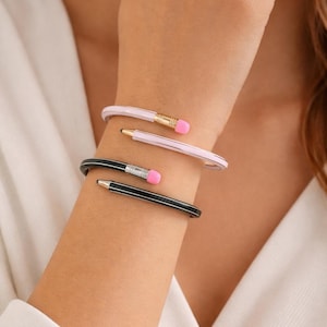 Pode incluir: São mostradas três pulseiras com tema de lápis. Duas são rosa claro e uma é preta. Cada pulseira tem uma faixa dourada ou prateada com uma borracha rosa e um design de ponta de lápis. As pulseiras são usadas no pulso.