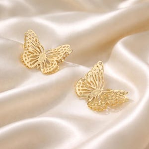 May include: Two gold-colored butterfly-shaped earrings. The earrings are made of a delicate, openwork design, with intricate details. They are set against a backdrop of a soft, cream-colored fabric.