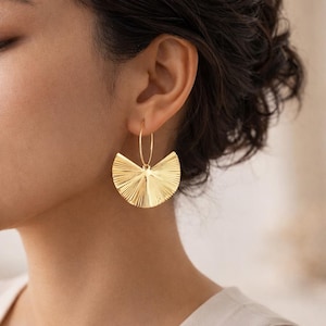 以下が含まれることがあります： 金色の扇形のイヤリング。イヤリングは、中心点から放射状に広がる、テクスチャ加工されたリブのデザインです。イヤリングは、細い円形のフープから吊り下げられています。イヤリングは明るく光沢のある金色です。