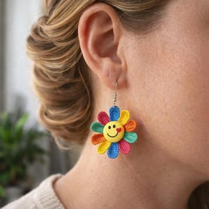 Pode incluir: Um brinco de margarida de crochê colorido com um rosto sorridente no centro. As pétalas são em tons de vermelho, laranja, amarelo, verde e azul. O brinco é preso a um gancho de prata.