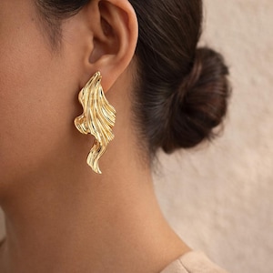 Peut inclure: Boucle d'oreille dorée unique, de forme abstraite et fluide. Le bijou présente une surface texturée et une forme organique et ondulée. Il s'agit d'une pièce de caractère, probablement en métal.