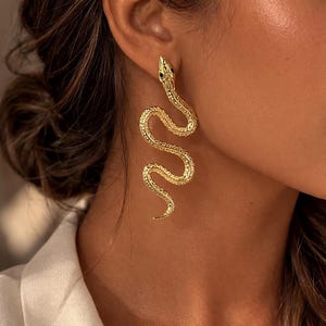 Può includere: Un lungo orecchino color oro a forma di serpente con una superficie testurizzata e squamosa e piccoli occhi neri. L'orecchino presenta un design sinuoso e serpentino che pende verticalmente, mostrando una finitura metallica dettagliata.