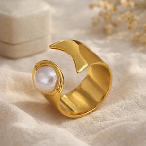 Peut inclure: Une bague dorée avec une grosse perle blanche sertie dans un design incurvé. La bague a une finition lisse et polie et un style moderne et ouvert. La perle est ronde et a un aspect brillant. La bague est probablement en métal.