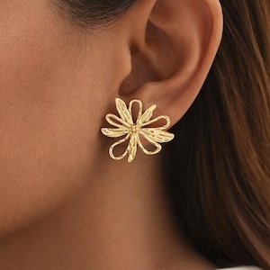 May include: A gold-colored flower-shaped stud earring. The earring is worn on the ear and has a delicate, openwork design. The petals are slightly curved, giving the earring a three-dimensional appearance. The earring is a bright, shiny gold color.