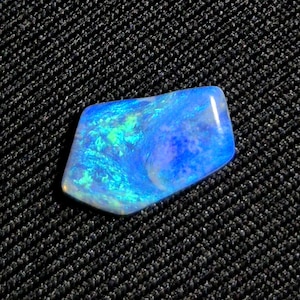 Könnte beinhalten: Ein polierter, schillernder Opal-Edelstein mit einer lebendigen Darstellung von Blau- und Grüntönen. Der Stein hat eine einzigartige, unregelmäßige Form und eine glänzende Oberfläche, die ein Farbspiel zeigt. Der Opal ist vor einem dunklen, strukturierten Hintergrund platziert.