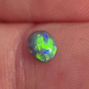 Pode incluir: Uma pequena pedra preciosa de opala negra de formato oval com uma cor de base escura e flashes iridescentes vibrantes de luz verde neon e azul brilhante que refletem em sua superfície polida, lisa e levemente arredondada.