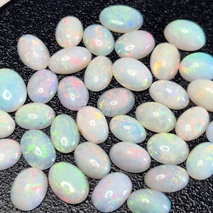 Pode incluir: Uma coleção de opalas ovais com uma cor de base branca leitosa. Cada pedra exibe flashes iridescentes de cor, incluindo rosa, azul e verde. As opalas estão espalhadas sobre uma superfície escura, mostrando sua beleza natural.