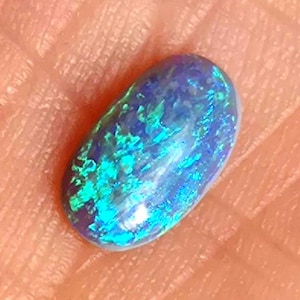 Könnte beinhalten: Ein ovaler Opal-Edelstein mit lebendigem Farbspiel. Der Stein hat eine tiefblaue Basis mit hellen türkisen und grünen Blitzen. Die polierte Oberfläche reflektiert das Licht und zeigt die inneren Farbvariationen des Steins.
