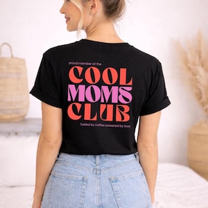 May include: Black t-shirt with the text "COOL MOMS CLUB" in red and pink lettering. The shirt also includes the phrases "proud member of the" and "fueled by coffee powered by love". The model is wearing light blue jeans.