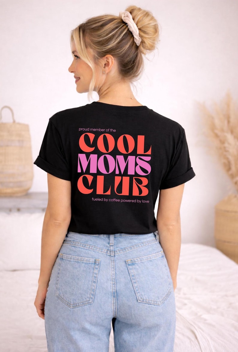 May include: Black t-shirt with the text "COOL MOMS CLUB" in red and pink lettering. The shirt also includes the phrases "proud member of the" and "fueled by coffee powered by love". The model is wearing light blue jeans.