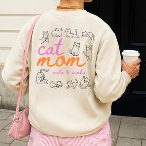 Könnte beinhalten: Cremefarbenes Sweatshirt mit dem Text "cat mom" in Rosa und Orange, umgeben von schwarzen Katzenillustrationen. Darunter steht "nala & simba". Die Person trägt rosa gestreifte Shorts und eine rosa Handtasche.