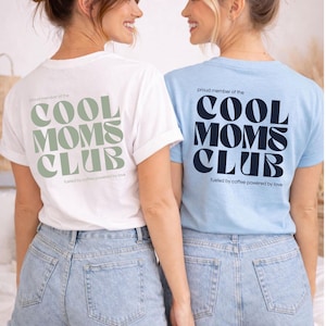 May include: Two women wearing white and light blue t-shirts with the text "COOL MOMS CLUB" in a bold font. The white shirt has green text, and the blue shirt has navy text. Both shirts have the phrase "fueled by coffee powered by love" below the club name.