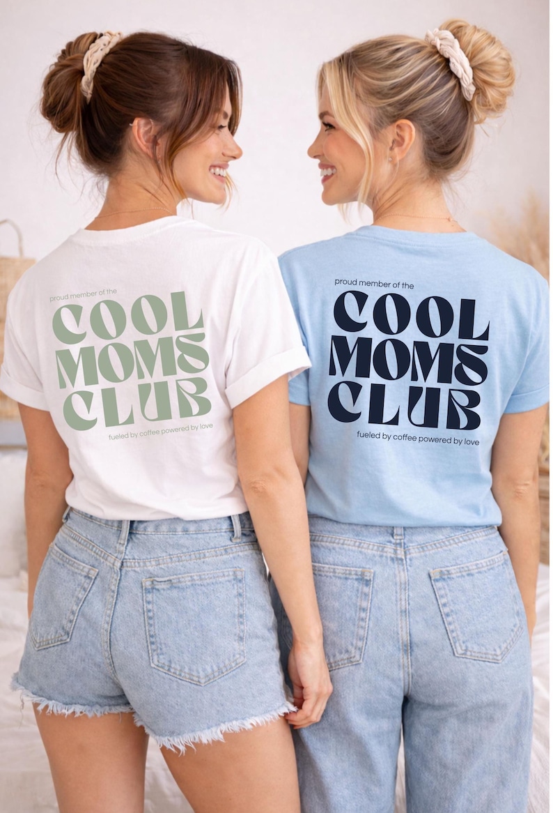 May include: Two women wearing white and light blue t-shirts with the text "COOL MOMS CLUB" in a bold font. The white shirt has green text, and the blue shirt has navy text. Both shirts have the phrase "fueled by coffee powered by love" below the club name.