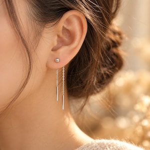 May include: A silver-toned earring featuring a small spherical stud connected to two delicate chains of varying lengths, each ending in a thin, straight metal bar. The earring is worn in the earlobe, with the chains hanging downward.
