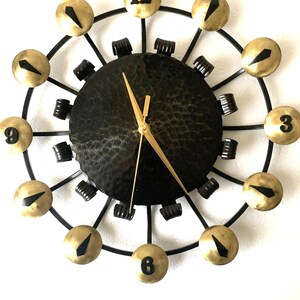May include: A decorative wall clock with a black hammered metal face and gold-coloured hands. The clock features gold-coloured spherical hour markers with black accents and a black metal frame. The clock is mounted on a white wall.