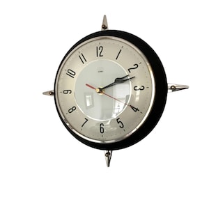 May include: A circular wall clock in a mid-century modern style, featuring a black frame and silver detailing. The clock face is off-white with black numerals and hands, plus a red second hand. It has four silver, pointed decorative elements.