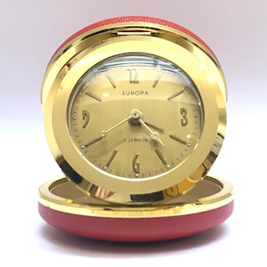 May include: A round Europa travel alarm clock with a gold-tone metal casing and a red textured exterior shell. The clock face is gold with black numerals at the 12, 3, 6, and 9 positions, featuring the text Europa and 2 Jewels in black print.