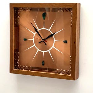 Puede incluir: Un reloj de pared cuadrado con marco de madera y esfera de color cobre. La esfera presenta un diseño de rayos de sol en blanco, manecillas negras para horas y minutos, un segundero rojo y cuatro detalles verdes en forma de gota.