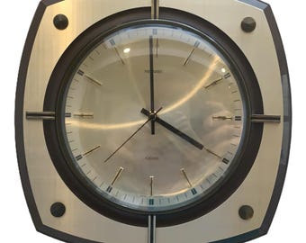 May include: A square wall clock with rounded corners, featuring a metallic gold face and a dark brown frame. The clock has thin black hands, gold hour markers, and four dark circular accents at the corners. The text "NIVAMEC" and "CHIME" are visible.