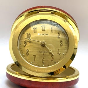 May include: A round Europa travel clock with a gold-toned brushed metal face, gold Arabic numerals, and three thin gold hands. The clock is encased in a polished gold metal frame set within a circular, reddish-brown textured base.