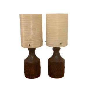 Puede incluir: Un par de lámparas de mesa con pantallas cilíndricas texturizadas en color blanco roto y bases de madera marrón oscuro con un patrón de veta visible. Cada lámpara tiene una pequeña fijación metálica que asegura la pantalla a la base.