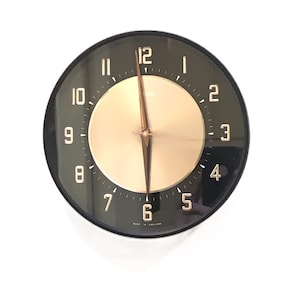 May include: A circular black wall clock with a gold-toned centre and gold hands. The clock face has white numerals and minute markers. The words "MADE IN ENGLAND" are printed at the bottom.
