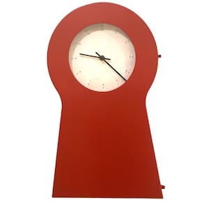 May include: A red clock with a keyhole-shaped frame and a circular white face. The clock features black hour, minute, and second hands, with small black numerical markings from one to twelve positioned around the perimeter of the white dial.