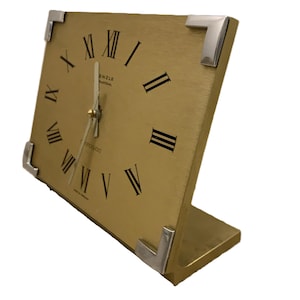 May include: A rectangular gold-toned desk clock with a brushed finish and silver-colored metal corner accents. The clock face features black Roman numerals, thin silver hands, and the text Kienzle International and Chronoquartz printed on it.