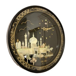 May include: Round black wall clock with a detailed mother-of-pearl inlay depicting a Middle Eastern cityscape. The clock face is black with gold-colored hands and hour markers. The outer rim features an ornate gold design. The clock measures approximately 12 inches in diameter.